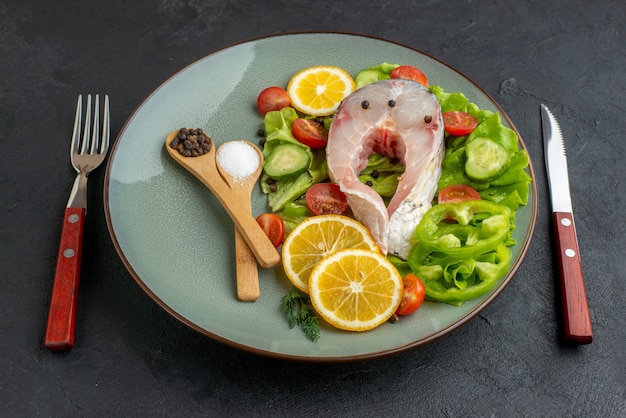 Vista laterale di pesce crudo e verdure fresche tritate fette di limone spezie su un piatto grigio e posate impostato su superficie nera in difficoltà