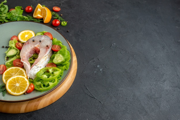 Vista laterale di pesce crudo e verdure fresche tritate fette di limone spezie su un piatto grigio su una tavola rotonda su superficie nera in difficoltà