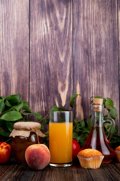 Vista laterale di pesche mature fresche con marmellata di pesche muffin in un barattolo di vetro e succo di pesca in un bicchiere su legno rustico