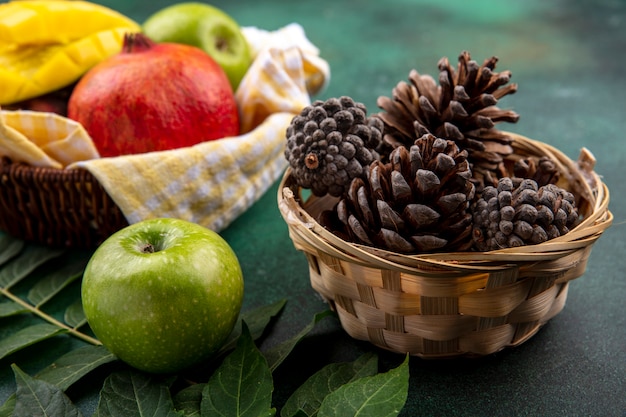 Vista laterale di pigne secche in un secchio con frutta fresca come melograno mango mela su busetto controllato giallo con foglia su superficie nera