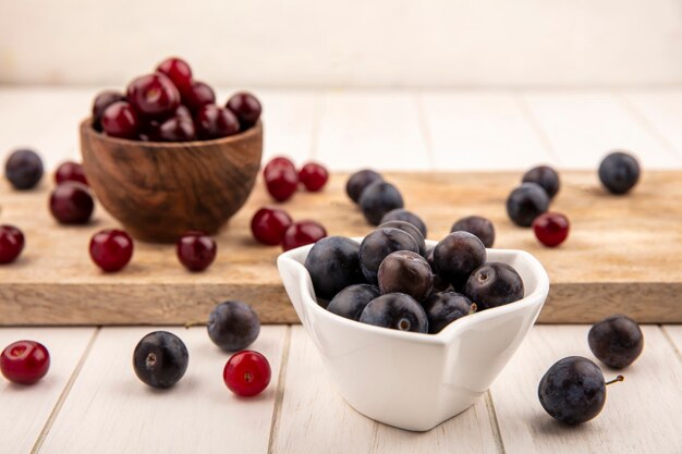 Vista laterale di prugnole viola scuro su una ciotola bianca con ciliegie rosse su una ciotola di legno su una tavola di cucina in legno su un fondo di legno bianco