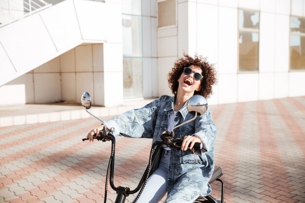 Vista laterale di risata della donna riccia in occhiali da sole che posano sulla motocicletta moderna all'aperto