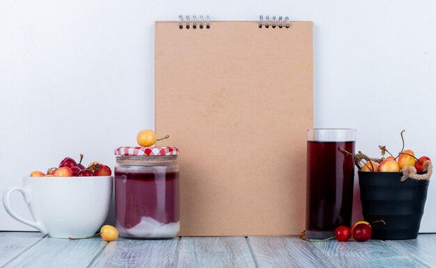 Vista laterale di sketchbook e ciliegie fresche più fresche in un secchio e ciotola bicchiere di succo e marmellata di ciliegie in un barattolo di vetro su rustico