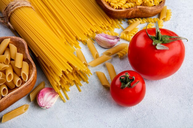 Vista laterale di spaghetti crudi con pasta cruda in ciotole con aglio e pomodori su una superficie grigia
