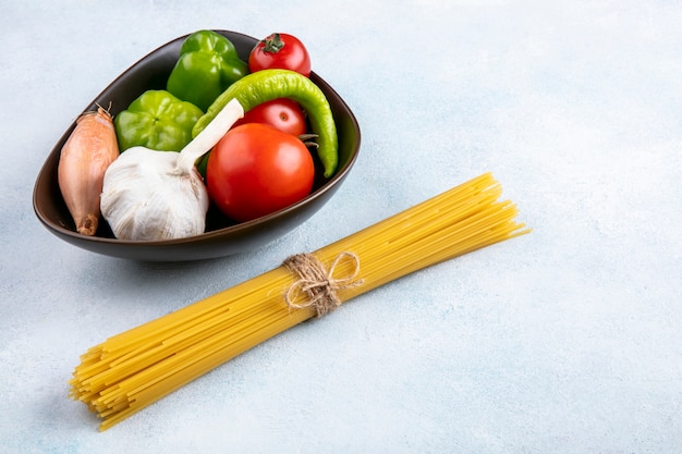 Vista laterale di spaghetti crudi con pomodori, aglio, peperone e cipolle in una ciotola su una superficie grigia