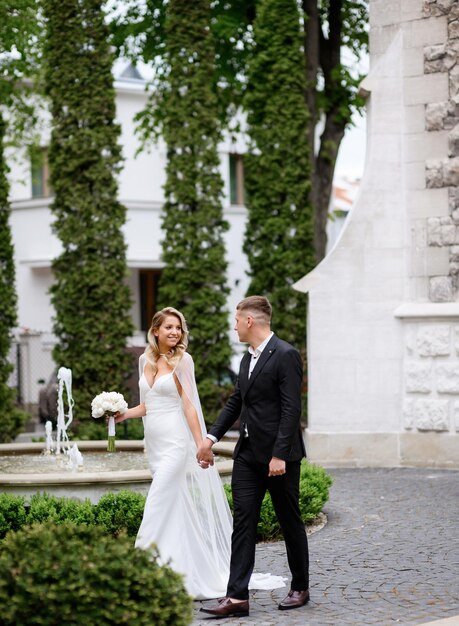 Vista laterale di sposi felici che indossano abiti da sposa tenendosi per mano guardandosi e godendosi l'atmosfera mentre camminano nel parco sullo sfondo di edifici e piante di architettura della fontana