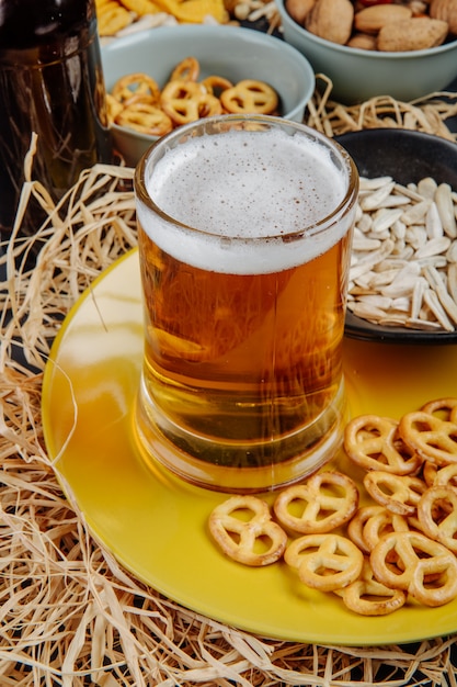 Vista laterale di un bicchiere di birra con mini salatini e semi di girasole sul piatto giallo su paglia