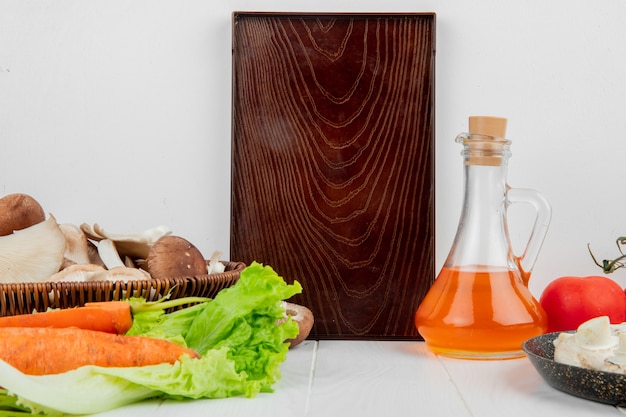 Vista laterale di un bordo di legno e dei funghi freschi in un canestro di vimini e nella bottiglia fresca delle carote di olio d'oliva su bianco