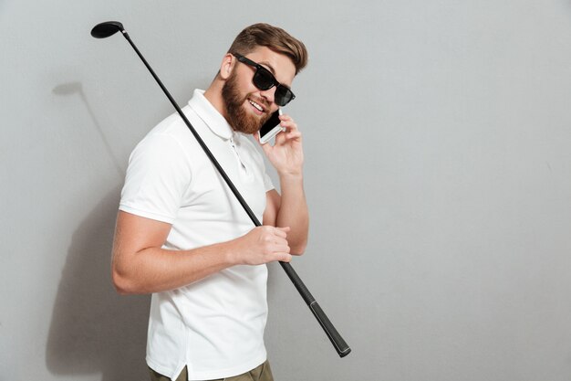 Vista laterale di un giocatore di golf calmo in occhiali da sole che parla dallo smartphone e che tiene club a disposizione sopra la parete grigia