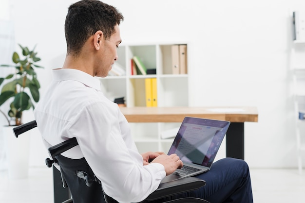Vista laterale di un giovane disabile che si siede sulla sedia a rotelle che utilizza computer portatile nell&#39;ufficio