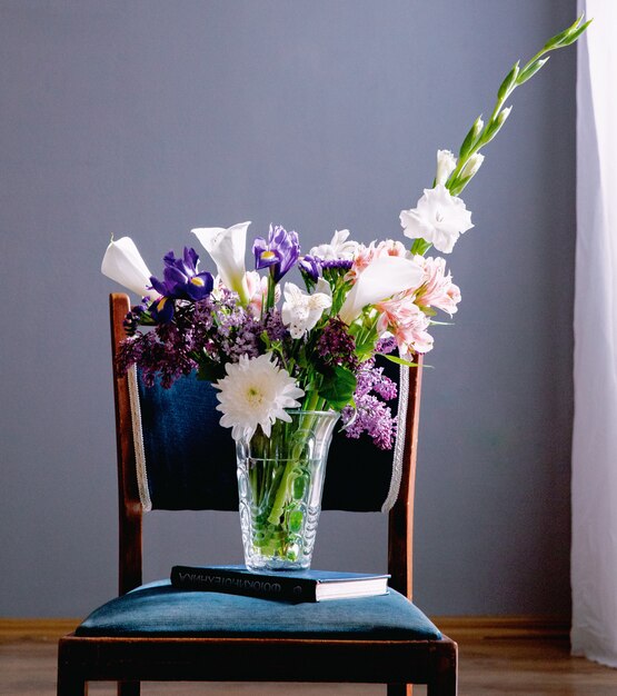 Vista laterale di un mazzo di calle di colore bianco con iris viola scuro lilla e fiori bianchi gladiolo in un vaso di vetro in piedi su un libro su una sedia a sfondo grigio muro