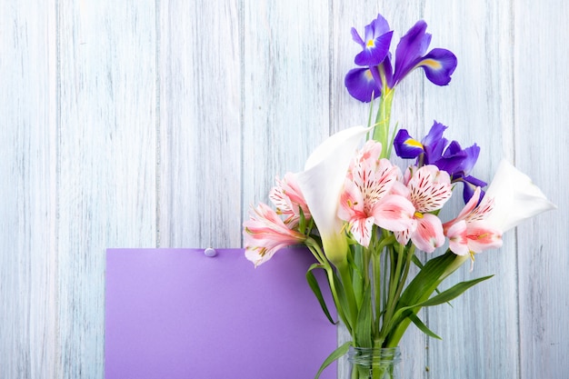 Vista laterale di un mazzo di fiori di colore rosa alstroemeria e fiori di iris viola scuro in una bottiglia di vetro con foglio di carta viola attaccato su fondo di legno grigio