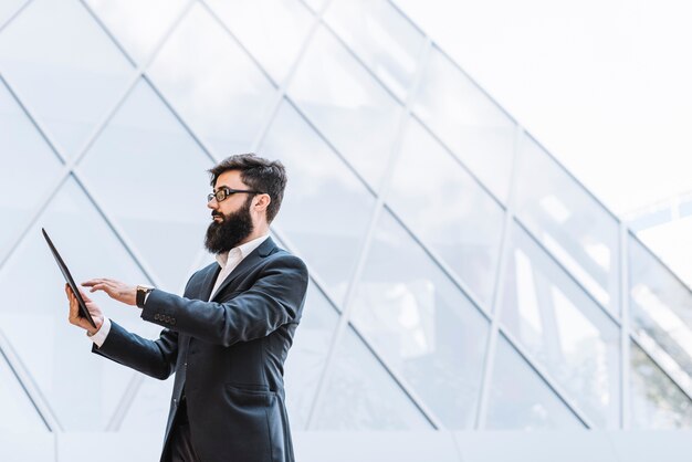 Vista laterale di un uomo d&#39;affari con la barba lunga che utilizza compressa digitale che sta contro la costruzione di vetro