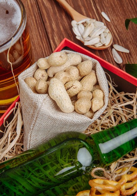 Vista laterale di una bottiglia di birra con le arachidi in un sacco in una scatola con paglia e una tazza di birra su rustico