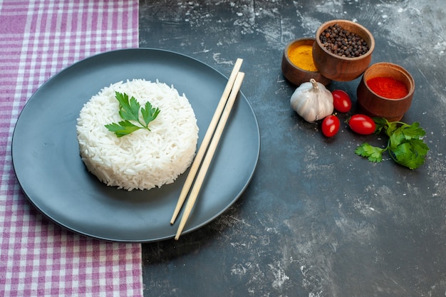Vista laterale di una deliziosa farina di riso servita con bacchette di legno verde su un piatto nero su un asciugamano spogliato viola e spezie diverse pomodori all'aglio su sfondo scuro