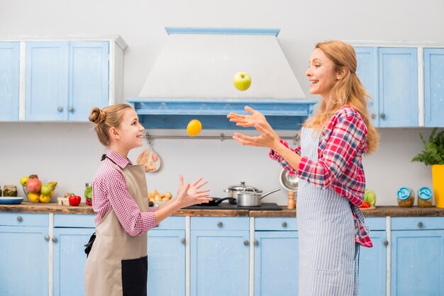 Vista laterale di una figlia e sua madre gettando mela e limone in aria in cucina