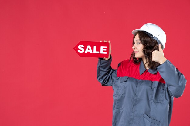 Vista laterale di una lavoratrice sorridente in uniforme che indossa elmetto e indica l'icona della vendita facendo un gesto ok su sfondo rosso isolato