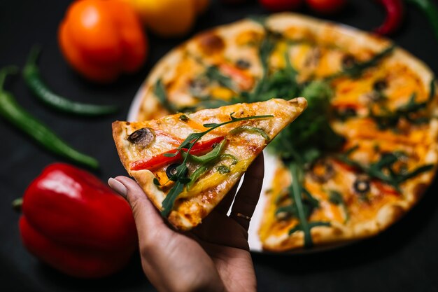 Vista laterale di una mano che tiene una fetta di pizza italiana con urugula e formaggio di olive nere di funghi peperoni colorati