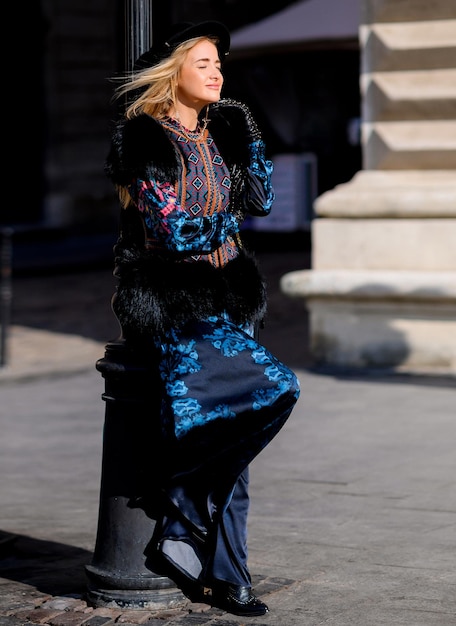 Vista laterale di una ragazza elegante e pensierosa che indossa un abito caldo e alla moda che alza la gamba e distoglie lo sguardo mentre si appoggia sulla lanterna di strada durante il freddo clima invernale