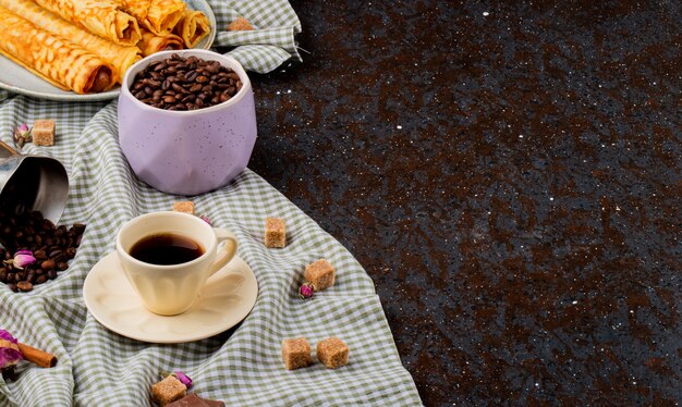 Vista laterale di una tazza di caffè e zucchero di canna cubetti di cioccolato e chicchi di caffè sparsi sulla tovaglia plaid con spazio di copia