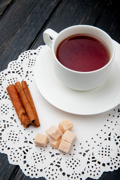 Vista laterale di una tazza di tè con bastoncini di cannella e cubetti di zucchero di canna sul tovagliolo di carta pizzo su fondo di legno scuro