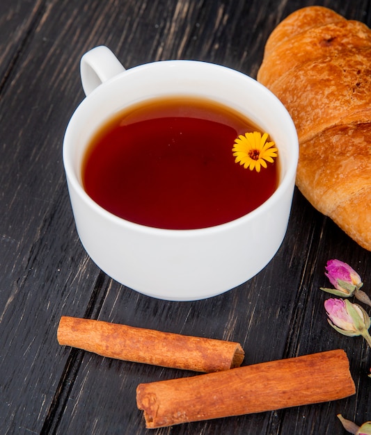 Vista laterale di una tazza di tè con croissant e bastoncini di cannella su legno nero