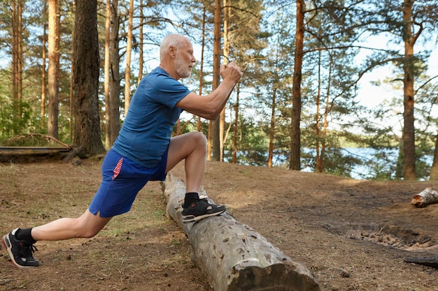 Vista laterale di uomo anziano forte in forma con la barba che lavora nella foresta, affondi doig, mantenendo i piedi sul registro Maschio anziano concentrato facendo esercizi fisici per i muscoli delle gambe in una giornata di sole estivo