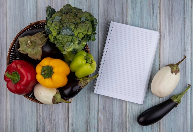 Vista laterale di verdure come broccoli pepe e melanzane nel cestino con blocco note su sfondo di legno con spazio di copia