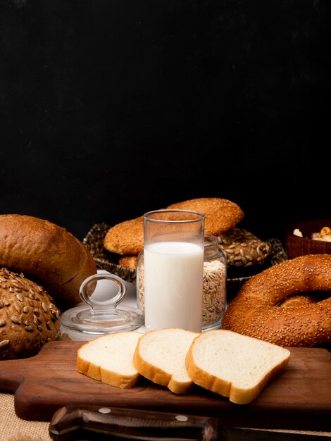 Vista laterale di vetro con latte e pane bianco affettato sul tagliere con il coltello e pani differenti su fondo nero con lo spazio della copia