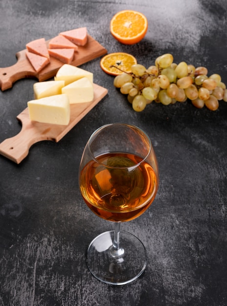 Vista laterale di vino bianco con l'uva, l'arancia ed il formaggio sul tagliere di legno sul verticale di superficie di buio