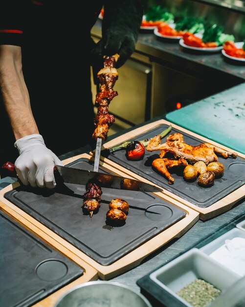 Vista laterale il cuoco rimuove da uno spiedino tike kebab su una tavola con pollo alla griglia e verdure