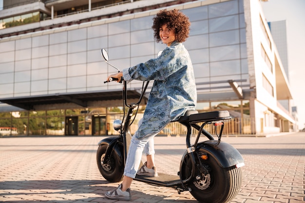 Vista laterale integrale della donna riccia sorridente che si siede sulla motocicletta