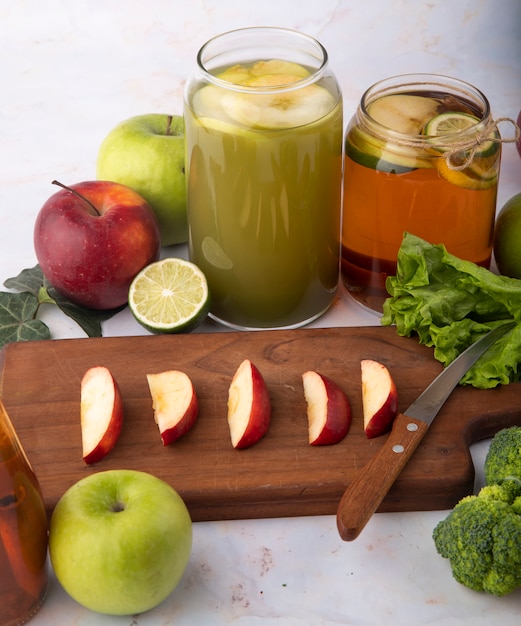 Vista laterale mix di mele succo di mela fresco brocoli tè al limone mela rossa affettata su una tavola mela verde fetta di lime e foglie di lattuga sulla superficie bianca