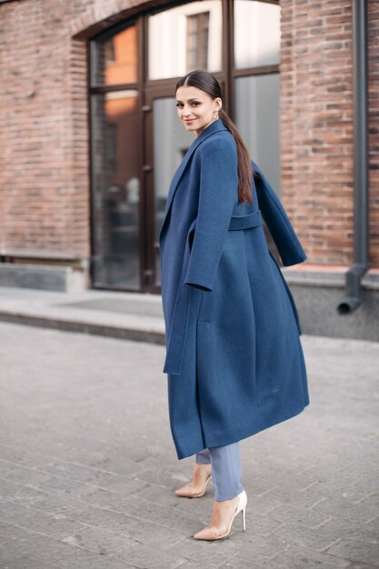 Vista laterale per tutta la lunghezza dell'attraente signora bruna con la coda che indossa cappotto blu, pantaloni azzurri e tacchi bianchi. Si sta muovendo e sorride alla telecamera in una strada urbana.