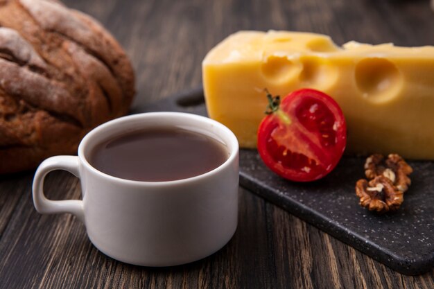 Vista laterale tazza di tè con formaggio maasdam e pomodoro su un supporto e pane nero sul tavolo