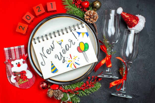 Vista orizzontale del taccuino con scritte e disegni di capodanno piatti per la cena accessori decorativi rami di abete calza di natale calici di vetro sul tavolo scuro