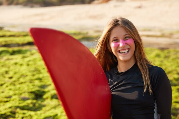 Vista orizzontale della donna attiva allegra con tavola da surf, sorride felicemente, tiene la tavola, posa all'aperto, vestita di nero