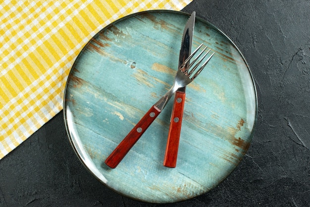 Vista orizzontale delle posate da pasto in croce su un piatto blu e asciugamano spogliato giallo su superficie scura dark