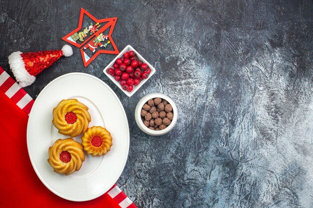 Vista orizzontale di deliziosi biscotti su un piatto bianco su asciugamano rosso e cappello di babbo natale cornel e chocoltes in vasi bianchi su superficie scura