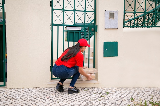 Vista posteriore del corriere femminile che mette i pacchi vicino al cancello. Deliverywoman bruna dai capelli lunghi in uniforme rossa accovacciata e consegna ordine espresso al cliente a casa. Servizio di consegna e concetto di posta