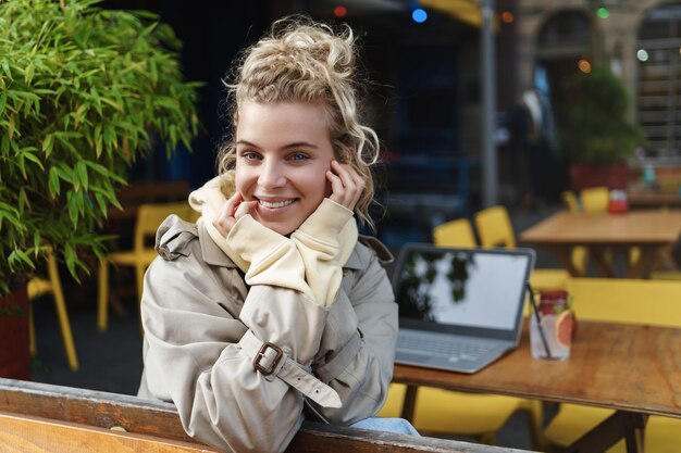 Vista posteriore del primo piano della bella ragazza bionda gira alla macchina fotografica mentre era seduto tavolino da caffè con computer portatile e cocktail.