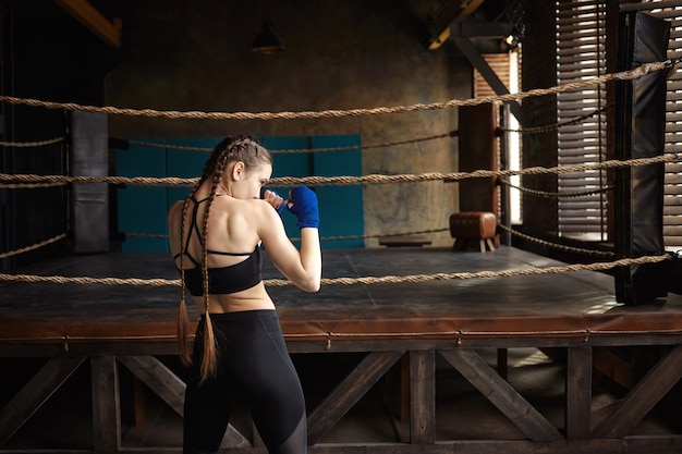 Vista posteriore del pugile professionista femmina con due trecce in piedi nella palestra vuota con ring di pugilato in background, formazione da solo,