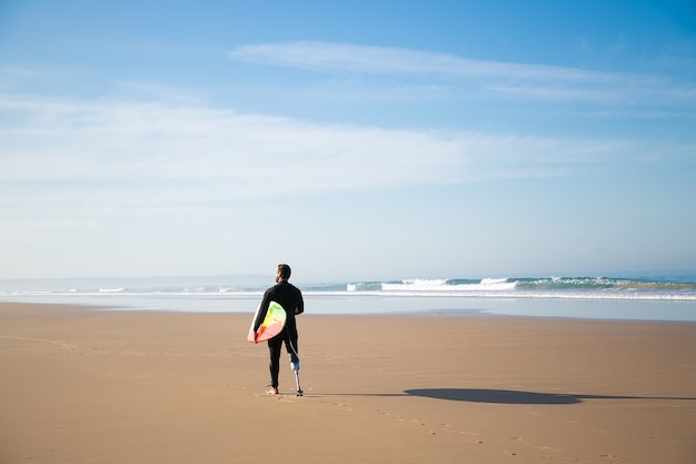 Vista posteriore del surfista in piedi sulla spiaggia di sabbia con bordo