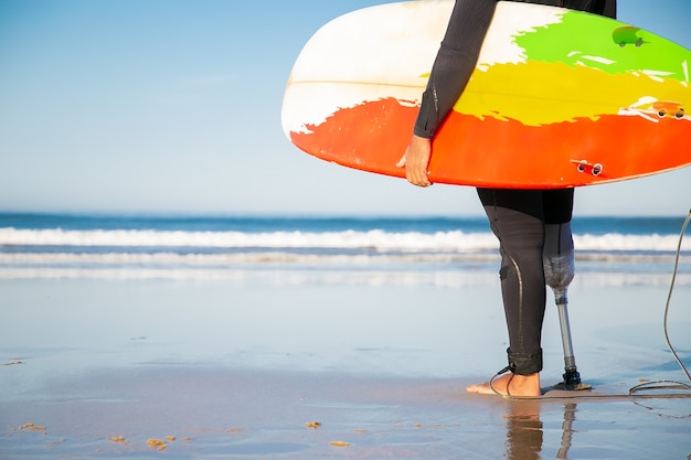 Vista posteriore del surfista maschio ritagliata in piedi con la tavola da surf sulla spiaggia del mare