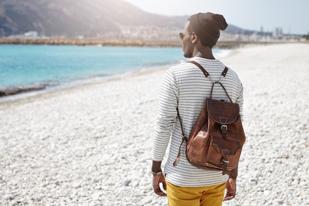 Vista posteriore del viaggiatore con zaino e sacco a pelo africano che affronta il mare in vestiti alla moda, viaggiando da solo nella località turistica europea, ammirando l'acqua e le montagne azzurro cielo, pensando a qualcosa di segreto e intimo