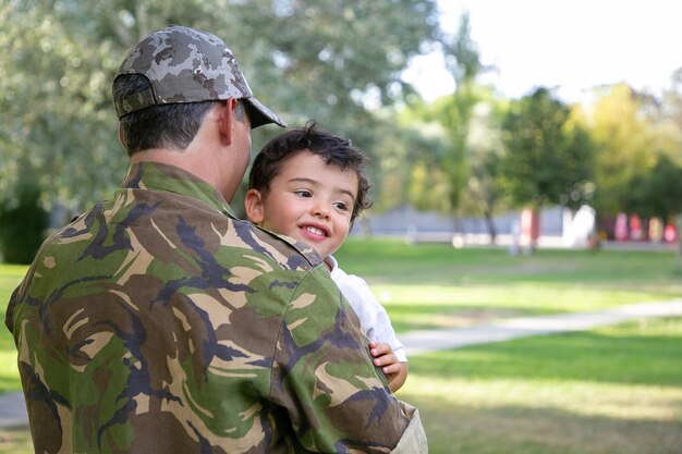 Vista posteriore dell'uomo caucasico che tiene il bambino e indossa l'uniforme dell'esercito. Ragazzino allegro che si siede sulle mani del padre, abbracciando il papà e sorridendo felicemente. Ricongiungimento familiare, paternità e concetto di ritorno a casa