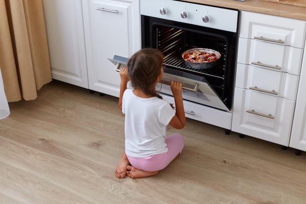 Vista posteriore della bambina in attesa di cuocere croissant, muffin o cupcakes vicino al forno, guardando dentro il forno mentre è seduta sul pavimento, bambina con le trecce che indossa una maglietta casual bianca.