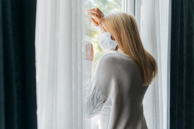 Vista posteriore della donna con mascherina medica a casa durante la pandemia guardando attraverso la finestra