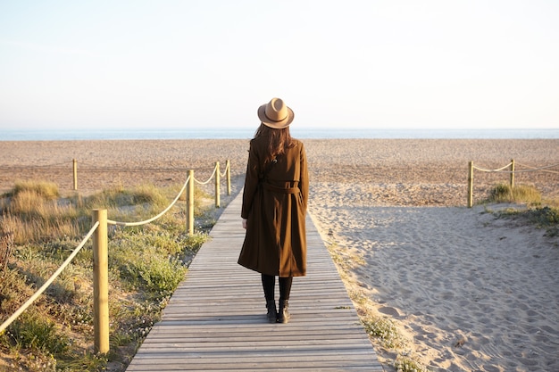 Vista posteriore della ragazza bruna in cappello e cappotto in piedi sul lungomare in riva al mare