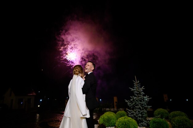 Vista posteriore della sposa senza volto vestita con abito bianco e giacca in piedi e guardando i petardi spettacolo in cielo mentre suo marito si sente felice e sorridente per strada durante la festa di matrimonio